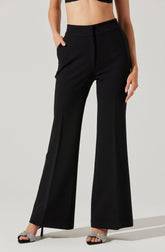 A woman is wearing high-waisted black wide-leg pants with a sleek, tailored appearance. The pants have a smooth waistband without visible belt loops and feature side pockets. She is also wearing a black cropped top, revealing her midriff, and silver high-heeled sandals with a sparkly finish.